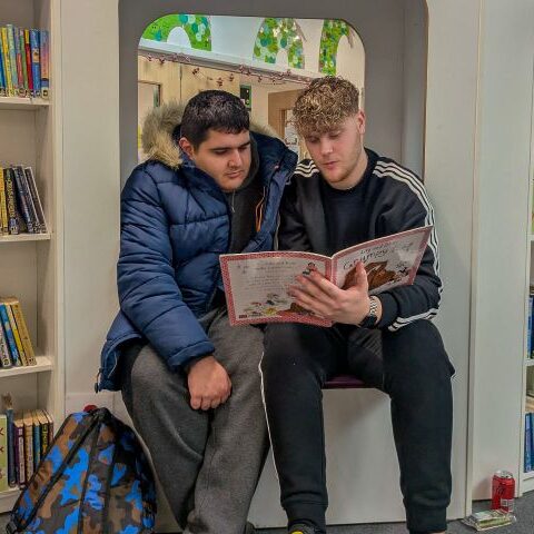 A Teens+ Project Worker reads a to a Teens+ student from a book in a library